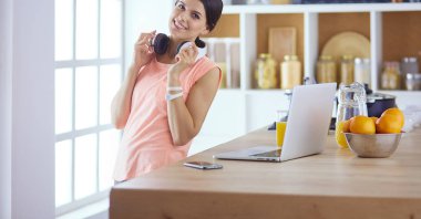Telefondaki güzel kadın evde, mutfakta dizüstü bilgisayar kullanıyor.