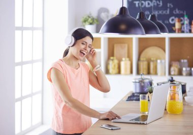Mutfakta dururken kulaklıkla müzik dinleyen ve dizüstü bilgisayar kullanan neşeli bir genç kadının portresi