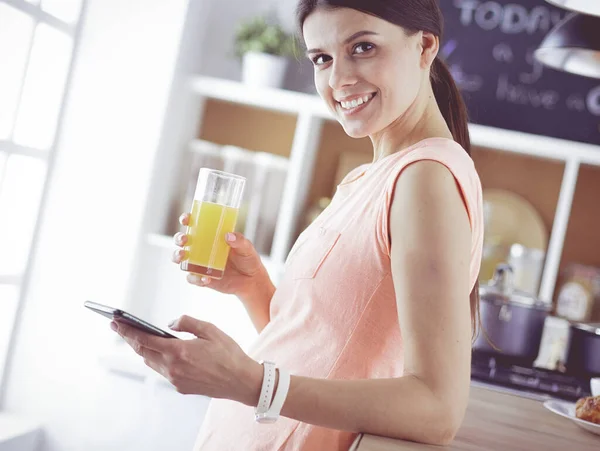 Leende vackra kvinna tittar på mobiltelefon och hålla glas apelsinjuice medan du äter frukost i ett kök. — Stockfoto