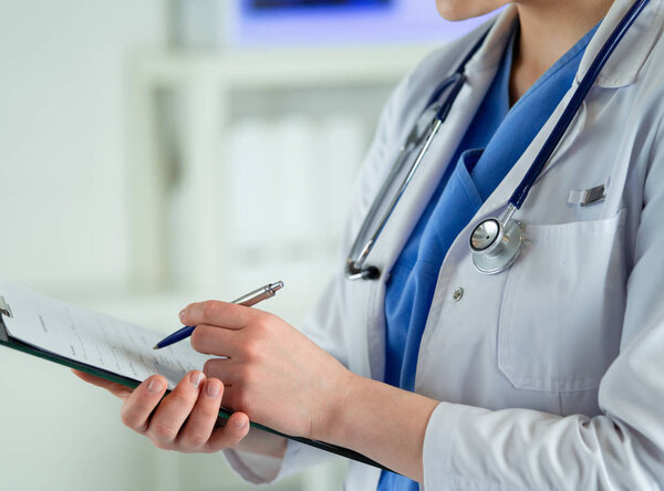 Doctor with a stethoscope, holding a notebook in his hand. Close-up of a female doctor filling up medical form at clipboard while standing straight in hospital