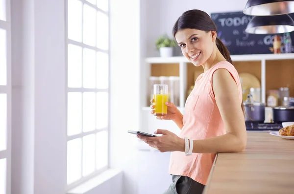 Mujer bonita sonriente mirando el teléfono móvil y sosteniendo un vaso de jugo de naranja mientras desayunaba en una cocina . — Foto de Stock