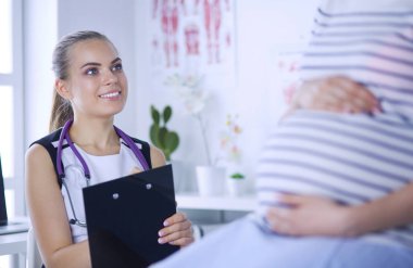 Steteskoplu ve tabletli genç kadın doktor hastanede hamile bir kadınla konuşuyor..