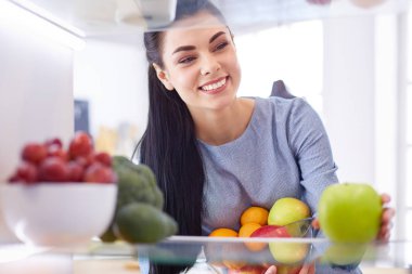 Gülümseyen kadın buzdolabından taze meyve alıyor, sağlıklı yiyecek konsepti.
