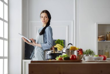 Mutfakta yemek yapmak için tablet bilgisayar kullanan genç bir kadın.