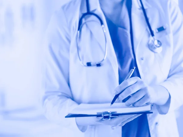 Doctor con un estetoscopio, sosteniendo un cuaderno en su mano. Primer plano de una doctora llenando el formulario médico en el portapapeles mientras está de pie en el hospital —  Fotos de Stock