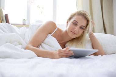 Elinde boş ekranlı dijital tablet tutan ve yatak odasında kameraya gülümseyen bir kız.