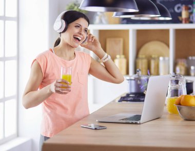 Mutfakta dururken kulaklıkla müzik dinleyen ve dizüstü bilgisayar kullanan neşeli bir genç kadının portresi