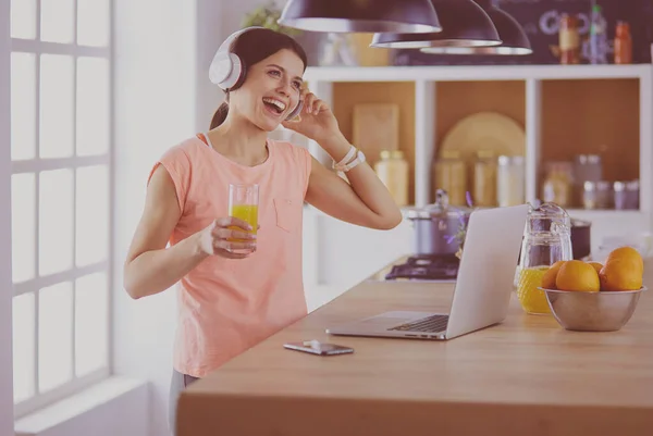 Porträtt av en glad ung kvinna lyssnar på musik med hörlurar och använda bärbar dator medan du står i köket — Stockfoto