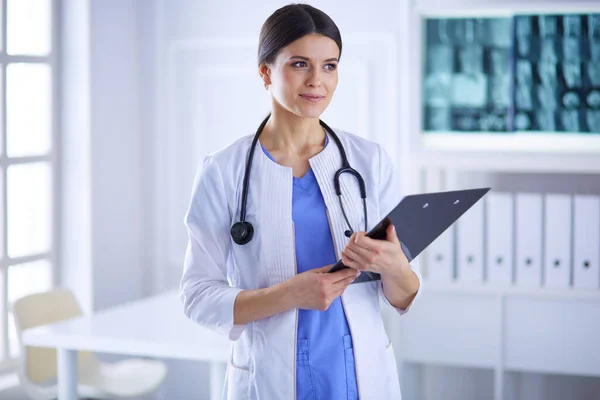 Joven doctora sonriente con estetoscopio sosteniendo una carpeta en el consultorio médico — Foto de Stock