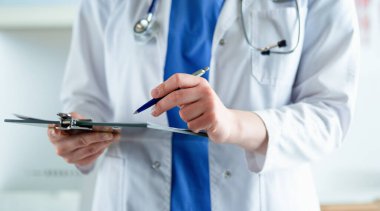 Steteskoplu bir doktor elinde bir defter tutuyor. Hastanede ayakta dikilirken panoda tıbbi form dolduran bir kadın doktorun yakın çekimi.