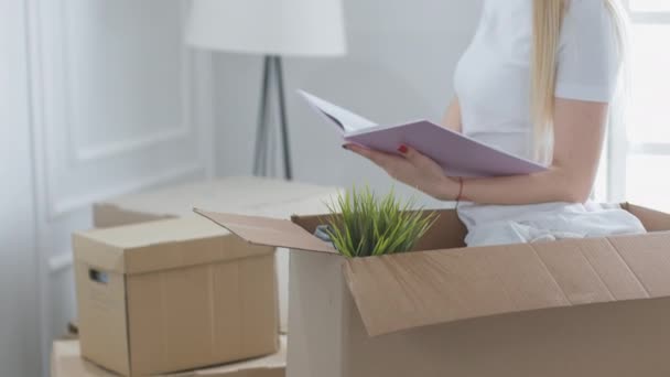Young woman moving into new place. Happy yong woman open unpacking cardboard box and holding a photo album — Stock Video