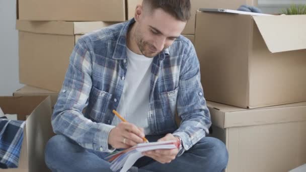 Un hombre en movimiento sentado en el piso en un apartamento vacío, Entre las Cajas, Comprobando la Lista de Cosas — Vídeos de Stock