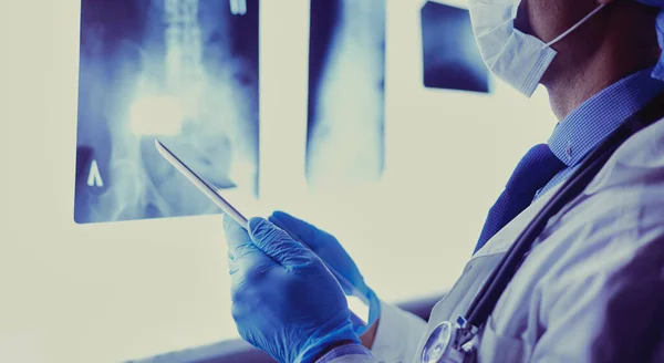 Arzt im Krankenhaus sitzt am Schreibtisch und betrachtet Röntgenbilder auf Tablet vor weißem Hintergrund mit Röntgenaufnahmen — Stockfoto