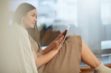 Evde oturan genç bir kadın pencerenin önünde oturuyor ve oturma odasında tablet bilgisayar kullanarak rahatlıyor.