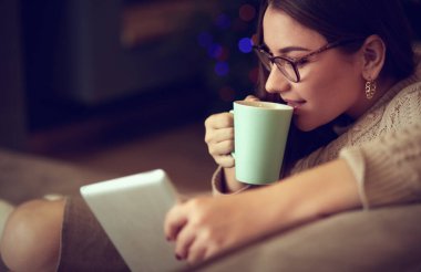Genç kadın evinde kanepede kahve içerken tablet kullanıyor.