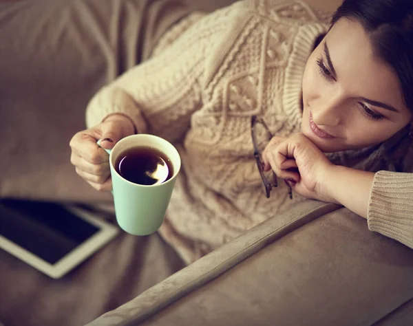 Kobieta w domu ubrania siedzi na dywanie w pobliżu kanapy za pomocą tabletu, pijąc kawę z zielonej filiżanki — Zdjęcie stockowe
