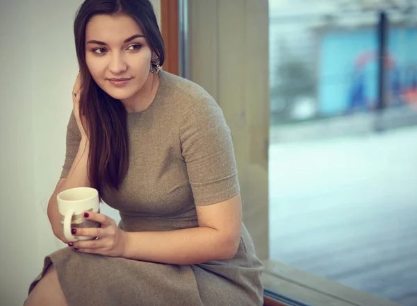 Jovem e bonita senhora sentada no peitoril da janela e olhando para fora da janela com xícara de café — Fotografia de Stock