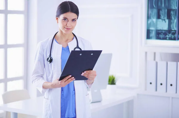 Joven doctora sonriente con estetoscopio sosteniendo una carpeta en el consultorio médico — Foto de Stock