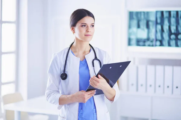 Joven doctora sonriente con estetoscopio sosteniendo una carpeta en una sala de consulta de hospitales — Foto de Stock