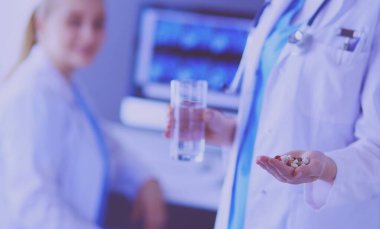 Doktorun arka planda meslektaşıyla klinikte elinde haplar ve bir bardak su tutarken görüntüsü..