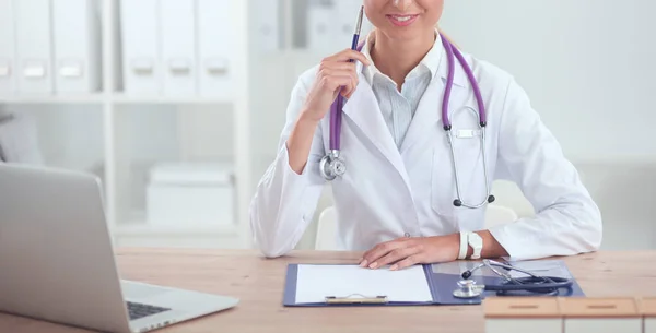 Bonito jovem sorridente médico feminino sentado na mesa e escrevendo. — Fotografia de Stock