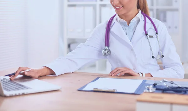 Bonito jovem sorridente médico feminino sentado na mesa e escrevendo. — Fotografia de Stock