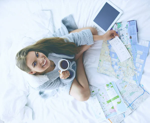 Relaxado jovem mulher sentada na cama com uma xícara de café e tablet digital — Fotografia de Stock