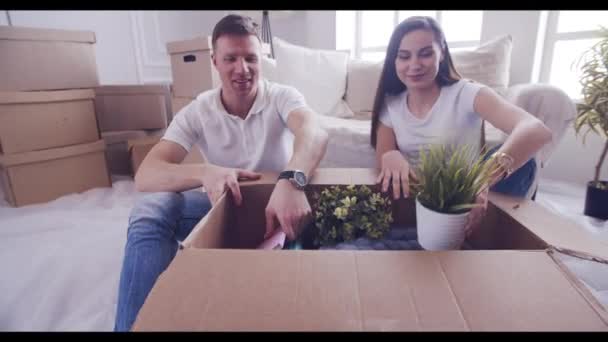 Emocionada pareja joven desempacando cosas, abriendo la caja y hablando durante el traslado a — Vídeos de Stock