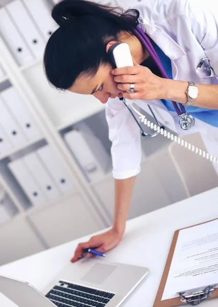 Young woman doctor in white coat at computer using phone — Stock Photo, Image