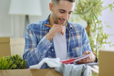 Taşınan bir adam boş bir dairede oturuyor, Kutuların arasında, eşyaların listesini kontrol ediyor.