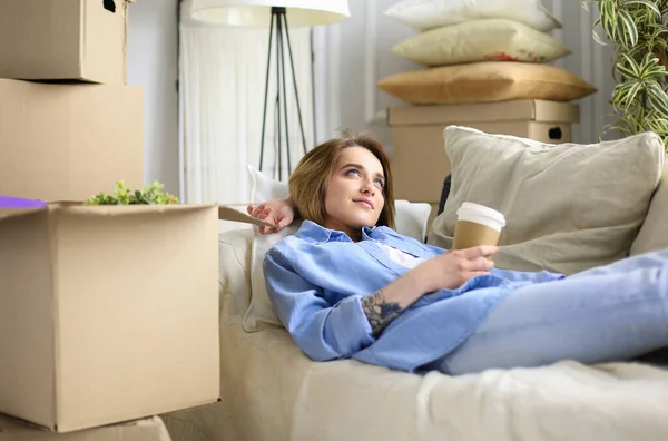 Счастливая молодая женщина лежит на диване в новом доме, пьет кофе или чай — стоковое фото