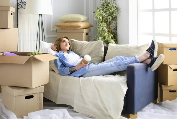 Ευτυχισμένη νεαρή γυναίκα ξαπλωμένη στον καναπέ στο νέο σπίτι, πίνοντας καφέ ή τσάι — Φωτογραφία Αρχείου