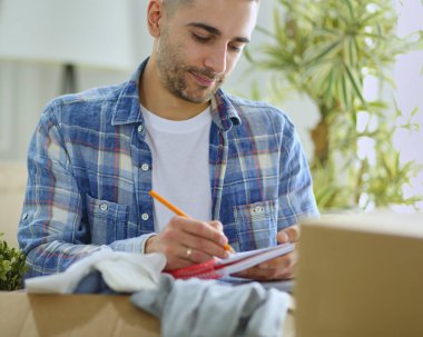 Taşınan bir adam boş bir dairede oturuyor, Kutuların arasında, eşyaların listesini kontrol ediyor.