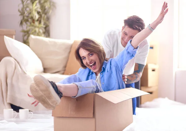 Casal se divertindo rindo movendo-se para a nova casa, jovem mulher montando sentado na caixa de papelão, enquanto o homem empurrá-lo — Fotografia de Stock