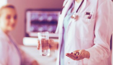 Doktorun arka planda meslektaşıyla klinikte elinde haplar ve bir bardak su tutarken görüntüsü..