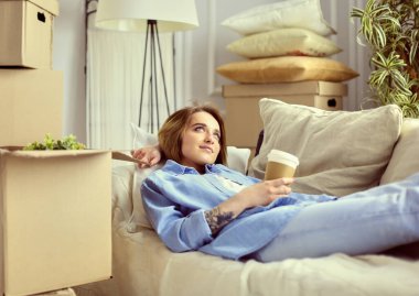 Yeni evindeki kanepede uzanmış, kahve ya da çay içen mutlu genç bir kadın.