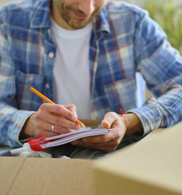 Taşınan bir adam boş bir dairede oturuyor, Kutuların arasında, eşyaların listesini kontrol ediyor.