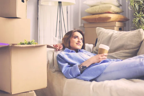 幸せな若い女性は新しい家でソファに横たわって、コーヒーや紅茶を飲んで — ストック写真