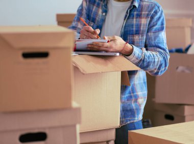 Taşınan bir adam boş bir dairede oturuyor, Kutuların arasında, eşyaların listesini kontrol ediyor.