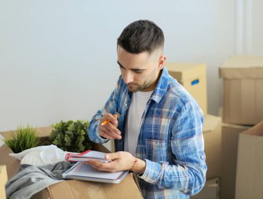 Taşınan bir adam boş bir dairede oturuyor, Kutuların arasında, eşyaların listesini kontrol ediyor.