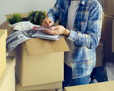 Taşınan bir adam boş bir dairede oturuyor, Kutuların arasında, eşyaların listesini kontrol ediyor.