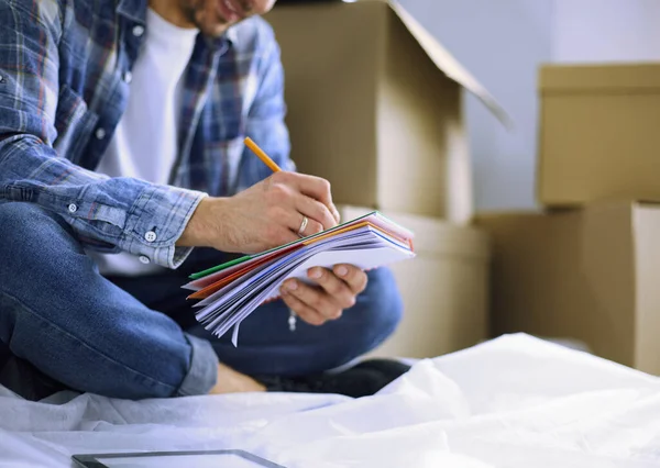 A moving man sitting on the floor in empty apartment, Among the Boxes, Checking the List of Things