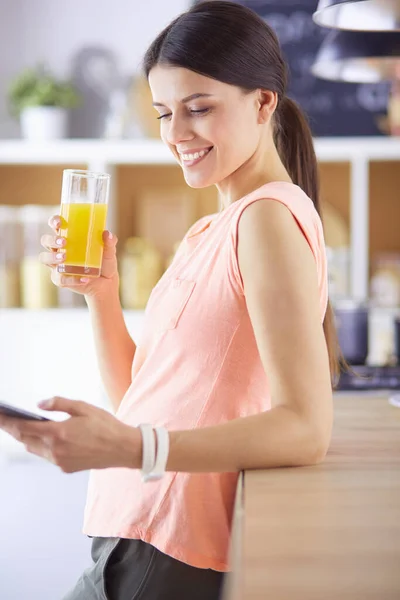 Smiling pretty woman looking at mobile phone and holding glass of orange juice while having breakfast in a kitchen. — Stock Photo, Image