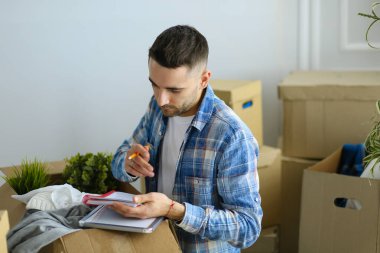 Taşınan bir adam boş bir dairede oturuyor, Kutuların arasında, eşyaların listesini kontrol ediyor.