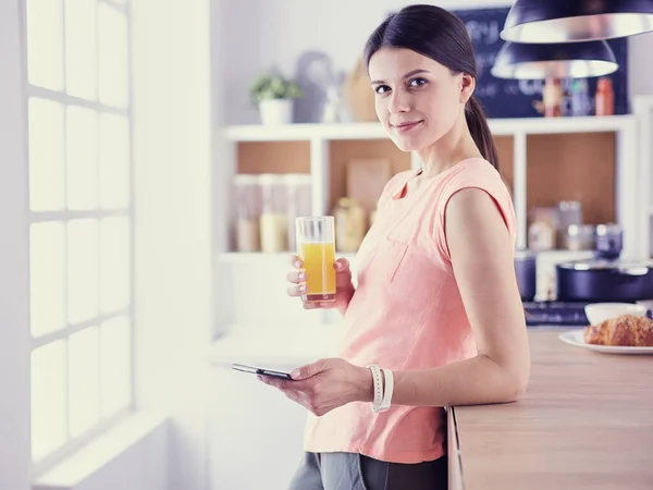 Leende vackra kvinna tittar på mobiltelefon och hålla glas apelsinjuice medan du äter frukost i ett kök. — Stockfoto