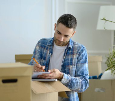 Taşınan bir adam boş bir dairede oturuyor, Kutuların arasında, eşyaların listesini kontrol ediyor.