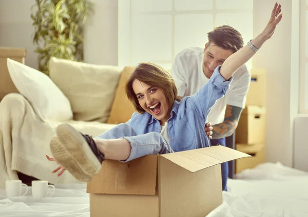 Casal se divertindo rindo movendo-se para a nova casa, jovem mulher montando sentado na caixa de papelão, enquanto o homem empurrá-lo — Fotografia de Stock