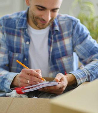 Taşınan bir adam boş bir dairede oturuyor, Kutuların arasında, eşyaların listesini kontrol ediyor.