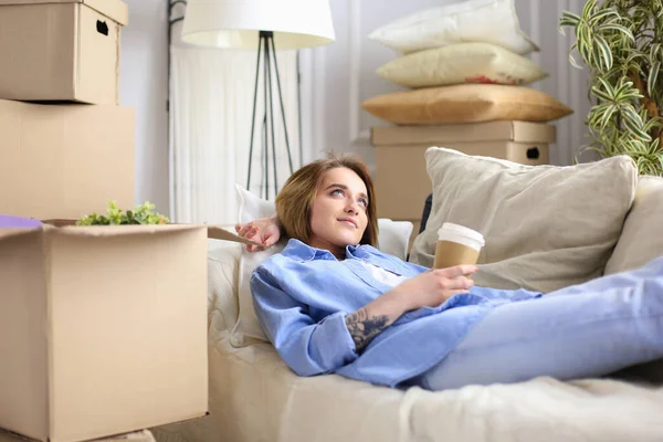 Lykkelig ung kvinde liggende på sofaen i nyt hjem, drikker kaffe eller te - Stock-foto