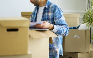 Taşınan bir adam boş bir dairede oturuyor, Kutuların arasında, eşyaların listesini kontrol ediyor.